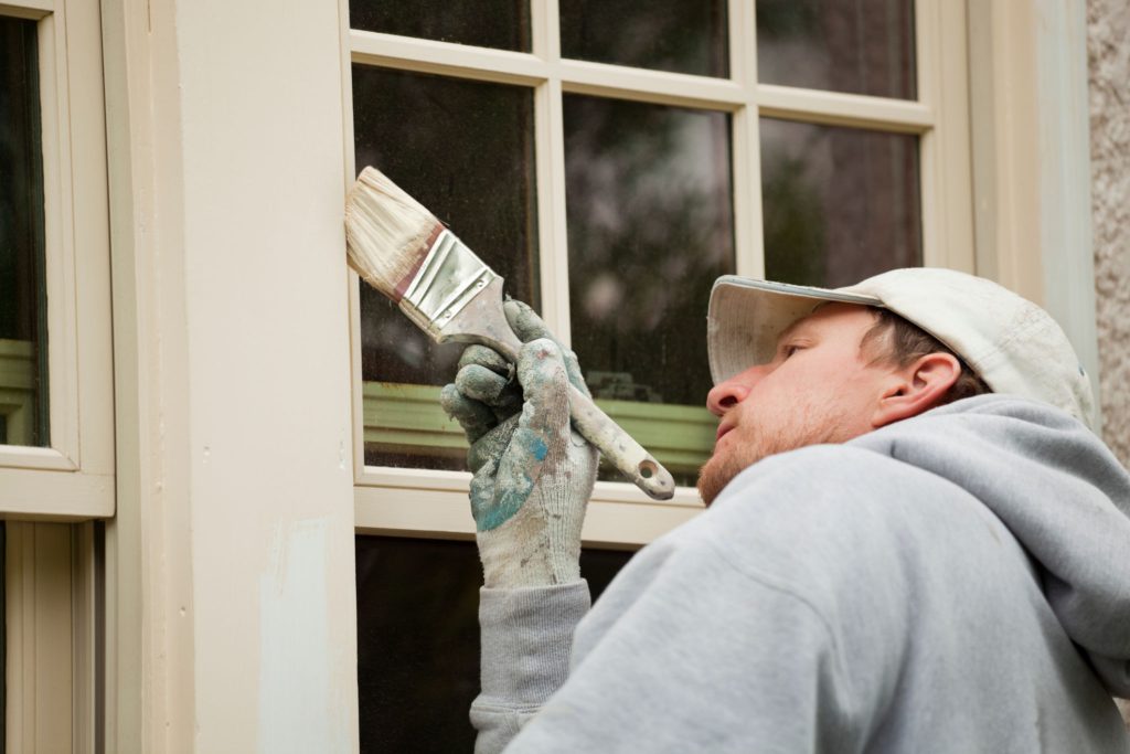 a man doing home maintenance, painting the window trim of a house.